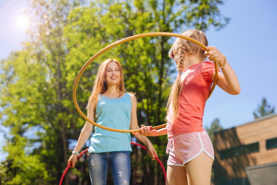 Pleasant interaction. Charming petite girl talking to her mother while hula-hooping in the back yard in the morning