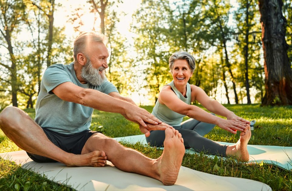 Sporty man and woman with grey hair stretching on yoga mats with hands to one leg during outdoors workout.