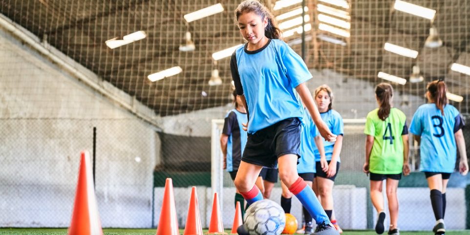 Young girl leading ball between cones on sports court. Female player in blue jersey practicing dribbling skill at court